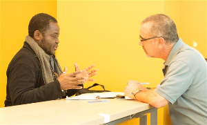 Two men dicussing as they sit at opposite sides of a long table. One is talking with his hands as the other listens intently. 