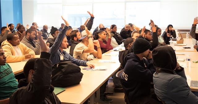 A large class of students, many of them have a hand raised.